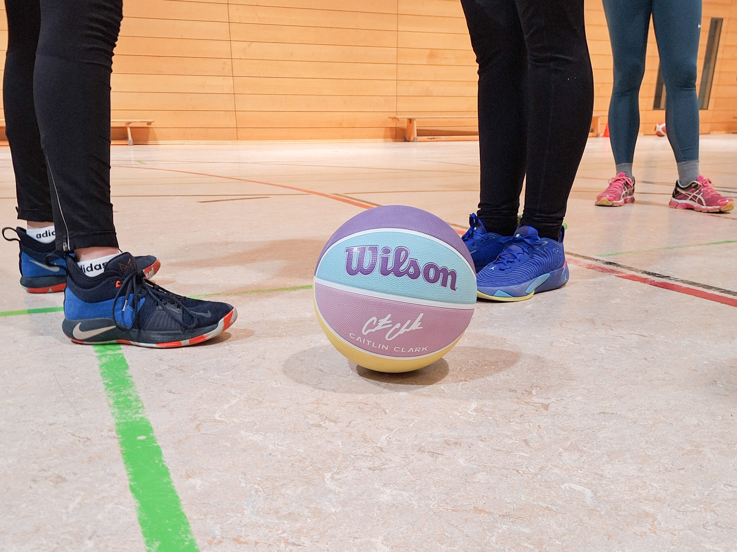 Lila-hellblau-rosa-farbener Basketball auf Turnhallenboden. im Hintergrund Füße in Basketballschuhen